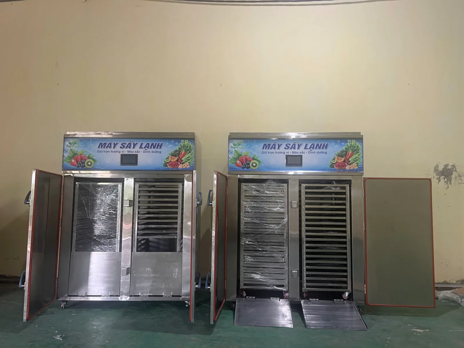 Hai máy sấy lạnh AGRITECH đặt cạnh nhau tại xưởng, cửa mở thấy giá khay inox